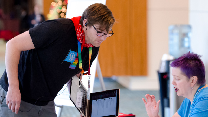 Two attendees discussing WordPress. Person on left is wearing a red bandana, a rainbow lanyard and precariously holding a laptop. The person on the right has deep purple hair and is wearing a light blue WordPress lanyard and t-shirt.