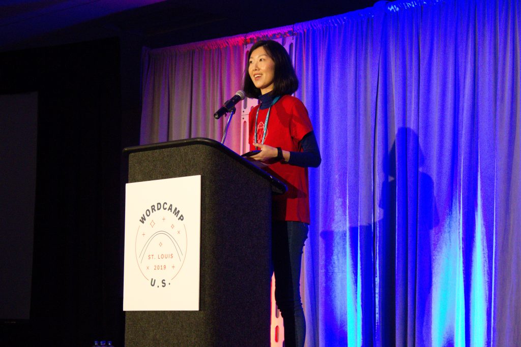 Woman in a red shirt with dark hair speaking behind a podium at WordCamp US 2019 in Saint Louis Photo Credit Ken Gagne