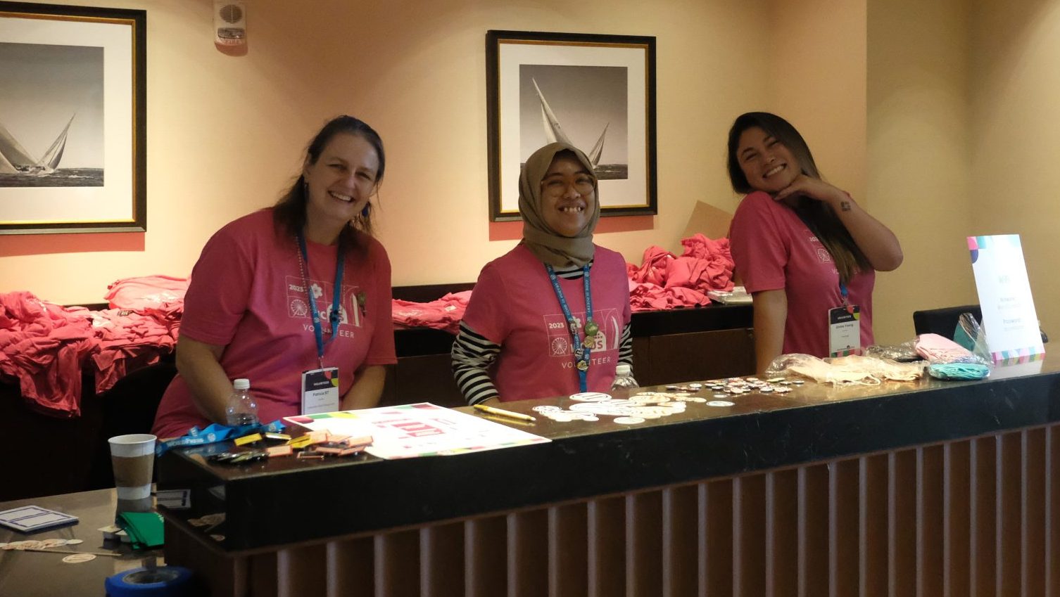 Three volunteers smile at the camera from behind the WordCamp registration desk.