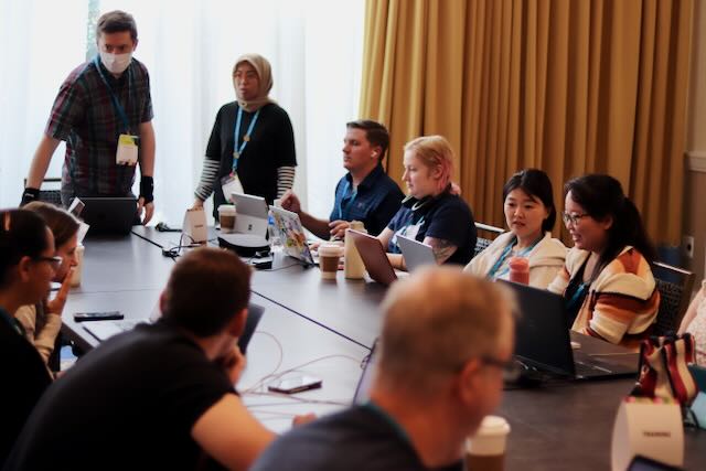 A diverse group of people from the WordPress Training Team is seated around a conference table at Contributor Day. Some are actively engaged in discussion, while others are focused on their laptops. A few participants are standing, including a person wearing a hijab and a person wearing a face mask. The atmosphere is collaborative, with participants of different backgrounds sharing ideas and working together. This image captures the spirit of teamwork and community involvement at WordCamp Contributor Day.